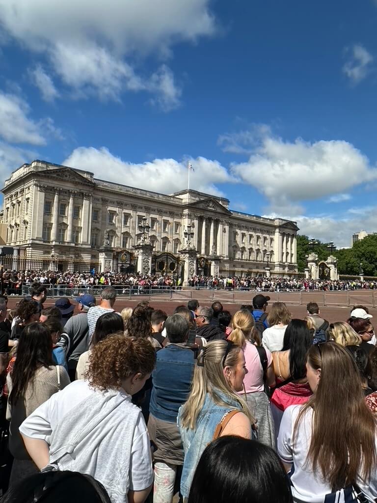 A crowd of people in front of Buckingham Palace on a blue skied sunny day with puffy white clouds.