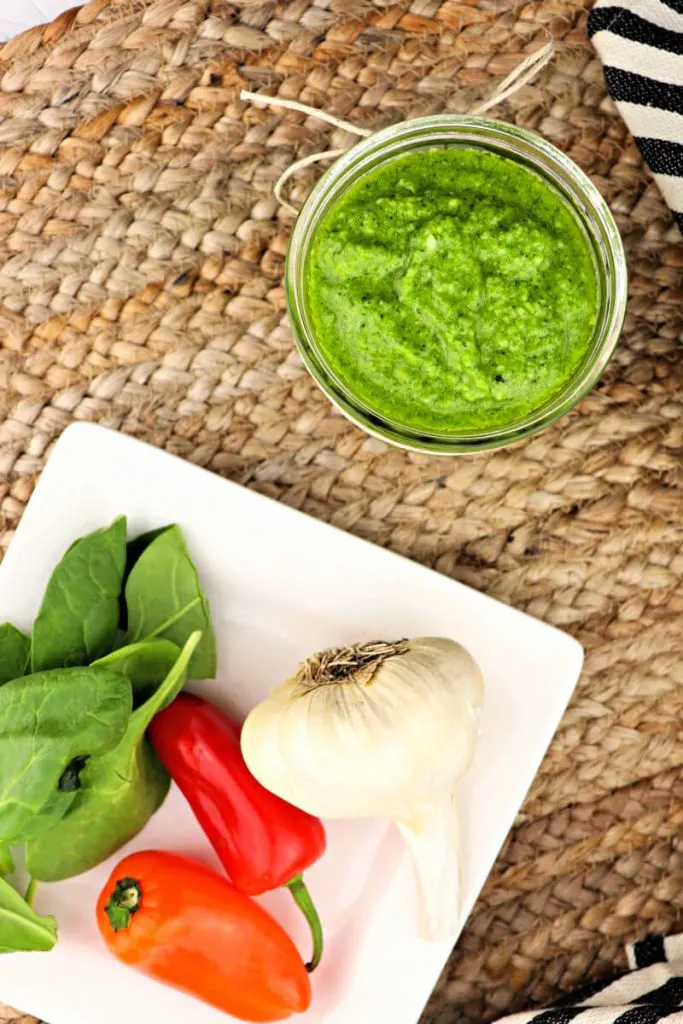 A jar of keto pest with a white plate of garlic, peppers, and spinach #ketorecipes #pesto