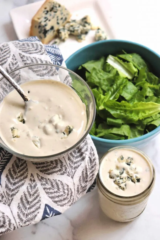Keto blue cheese dressing in a bowl and jar with a bowl of salad and blue cheese in back.
