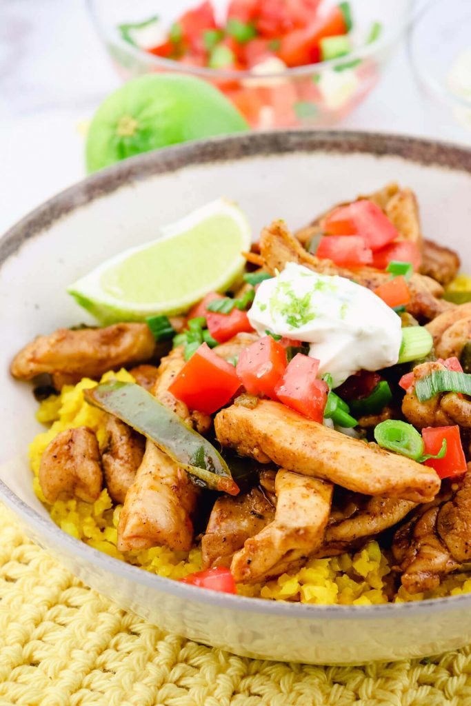 A bowl of taco chicken over cauliflower rice with pico de gallo, roasted peppers, sour cream, lime zest, etc. Sitting on a yellow crocheted mat.