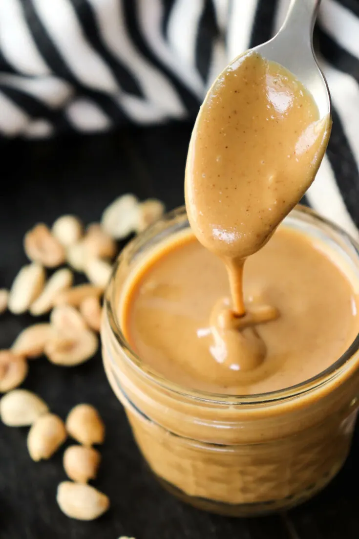 Spoon dripping peanut butter into a jar with peanuts on the table