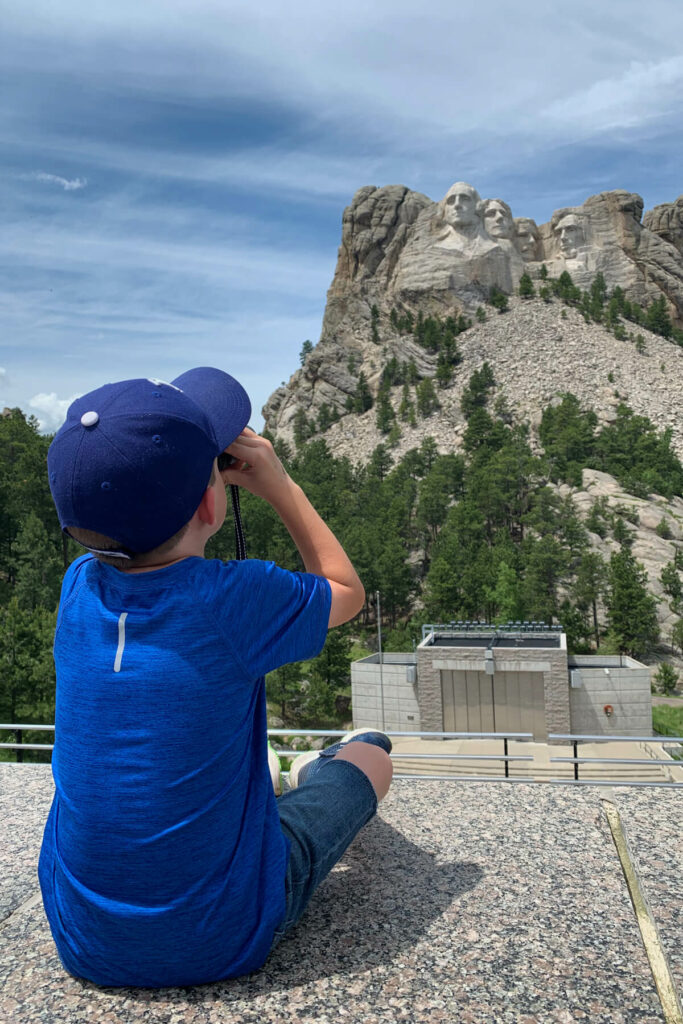 Boy in blue looking through binoculars towards Mount Rushmore