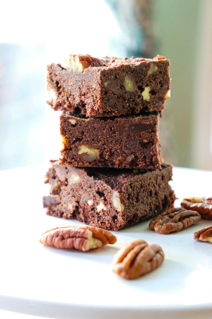 A stack of three yummy keto coconut flour brownies on a white platter with pecans