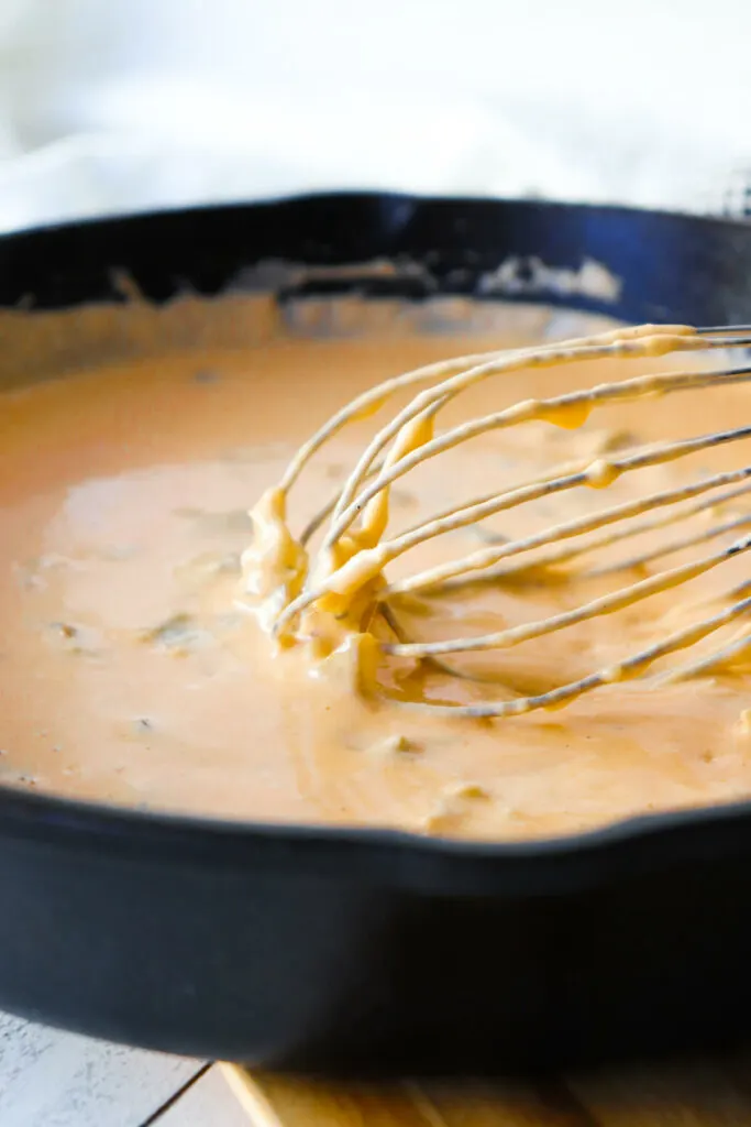 Front view of a whisk in green chile keto queso dip in a cast iron pan