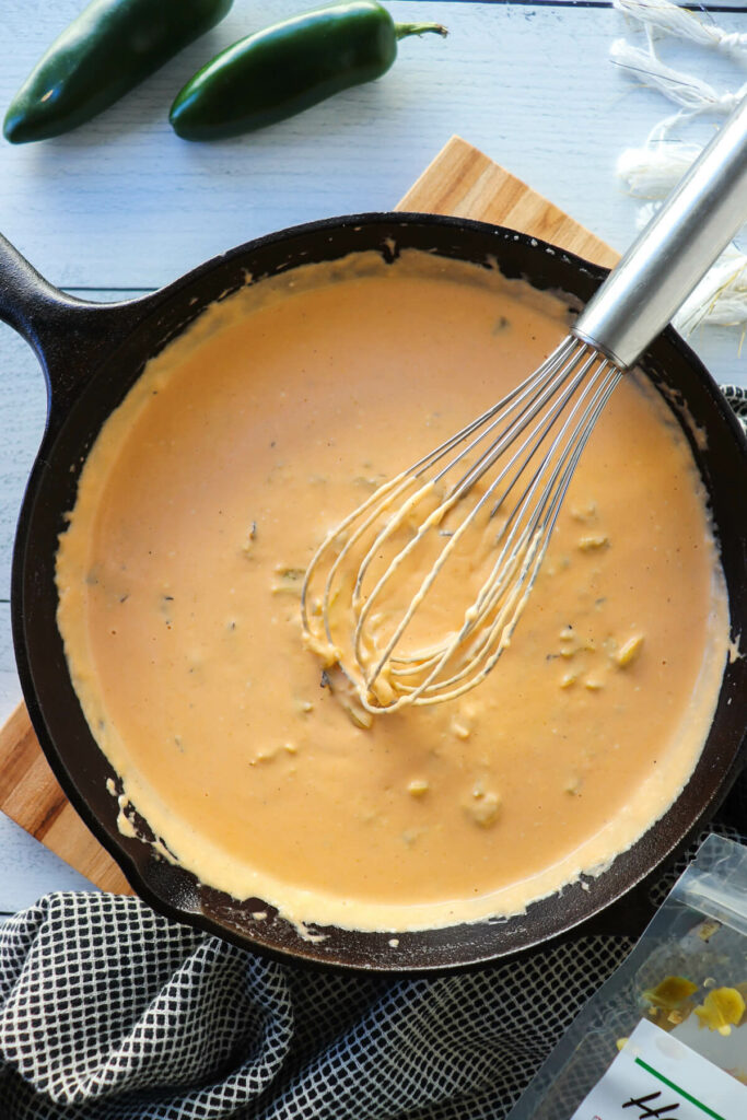 Overhead view of a whisk in green chile keto queso dip in a cast iron pan