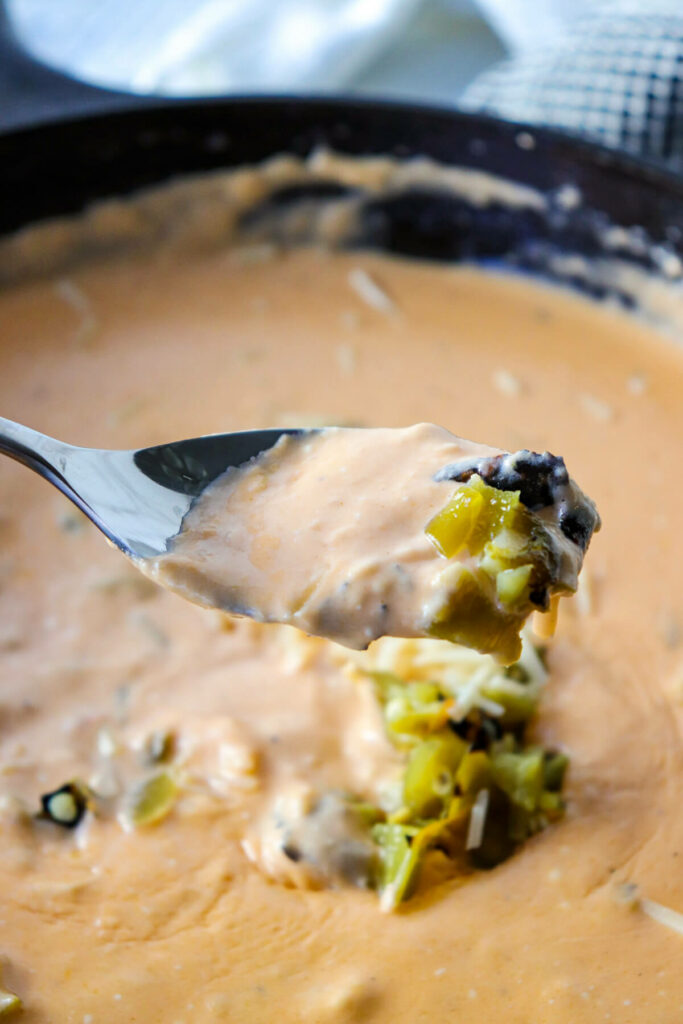 A spoon holding a bite of keto queso dip over a cast iron skillet of green chile cheese dip