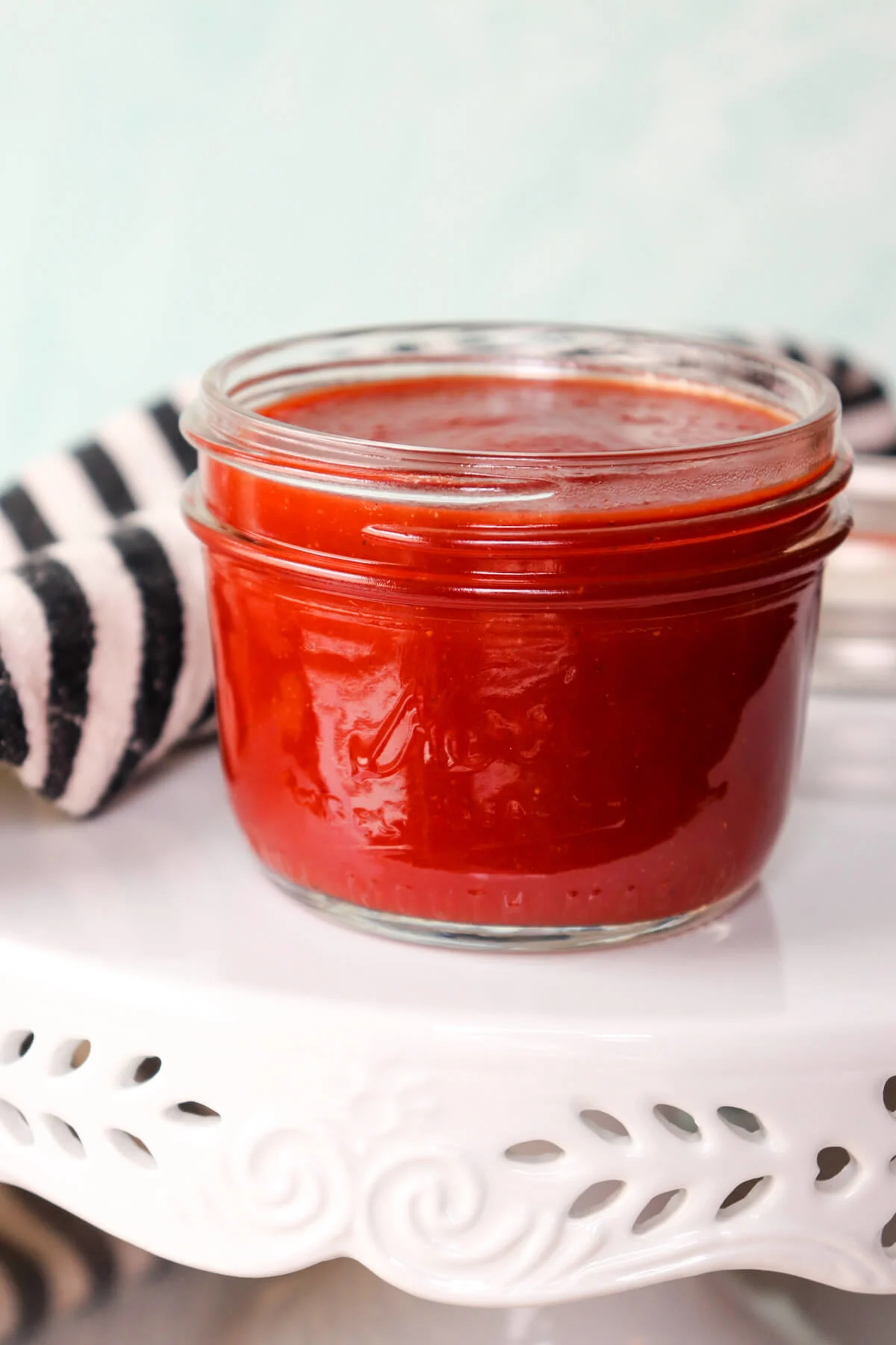 A glass jar of keto ketchup on a white stand with a black and white kitchen towel in the background.