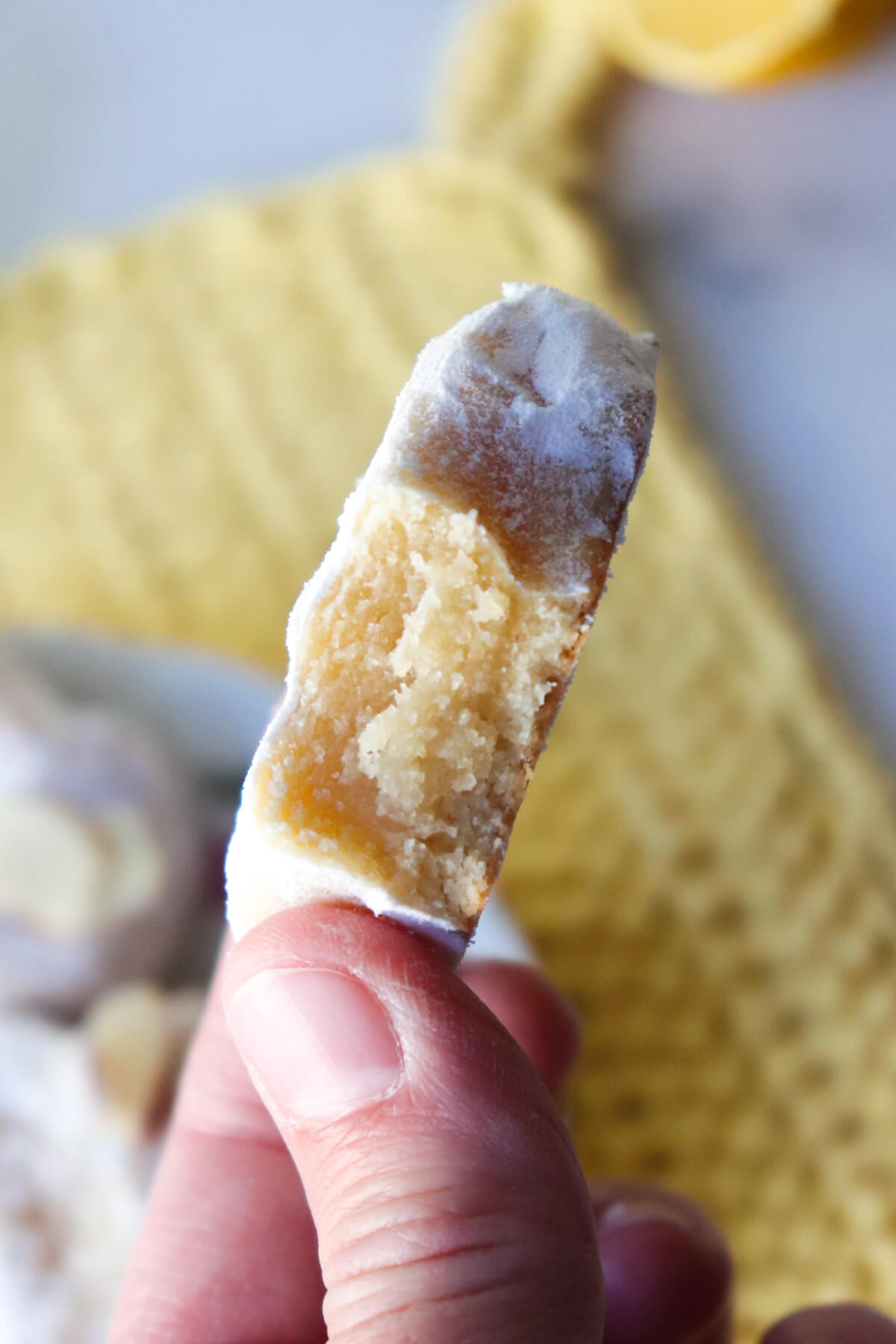 A bite out of a Greek almond cookie with a hand holding it up and a yellow kitchen cloth in the background