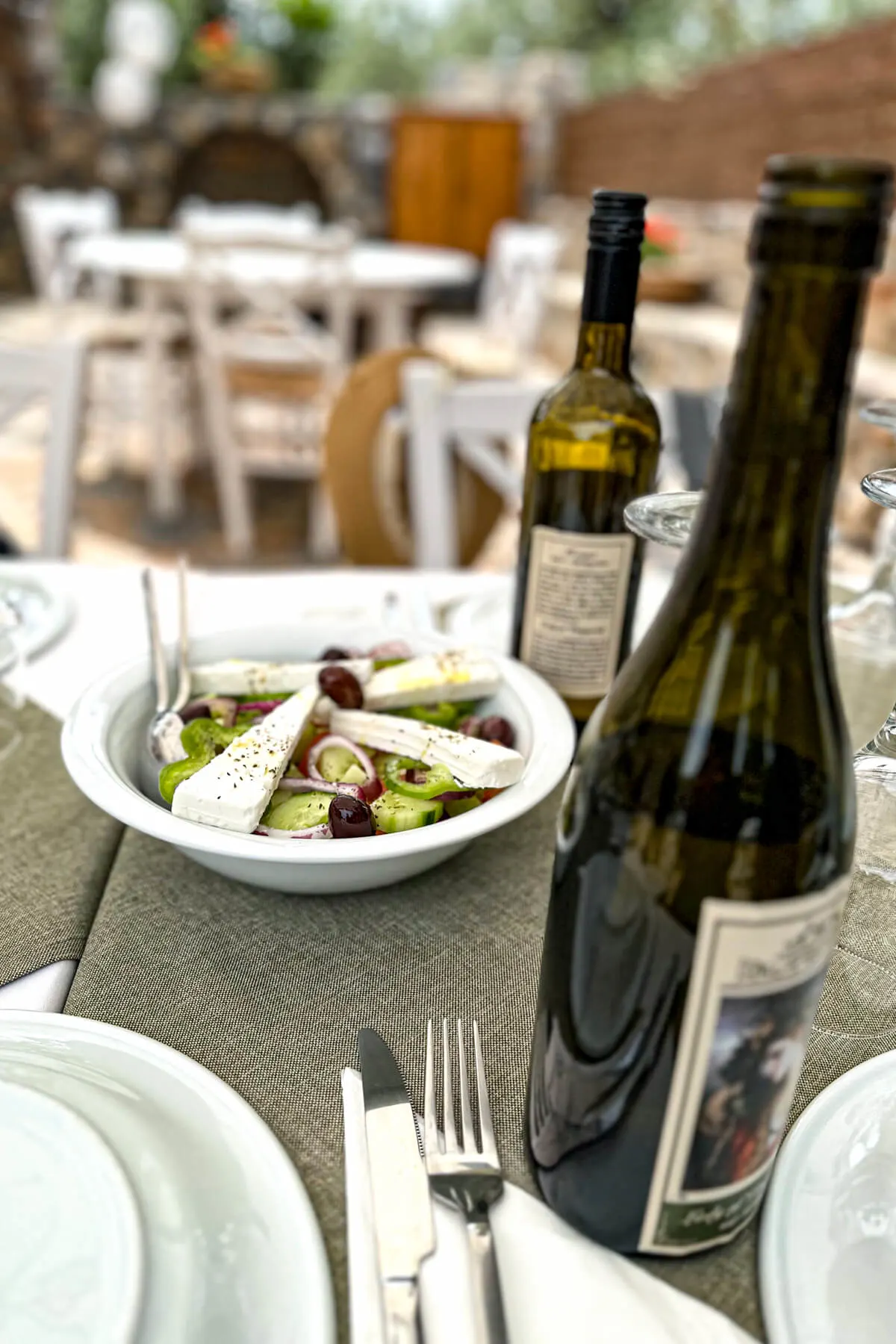 A Greek cucumber salad on a set table outdoors with wine bottles and tables in the background