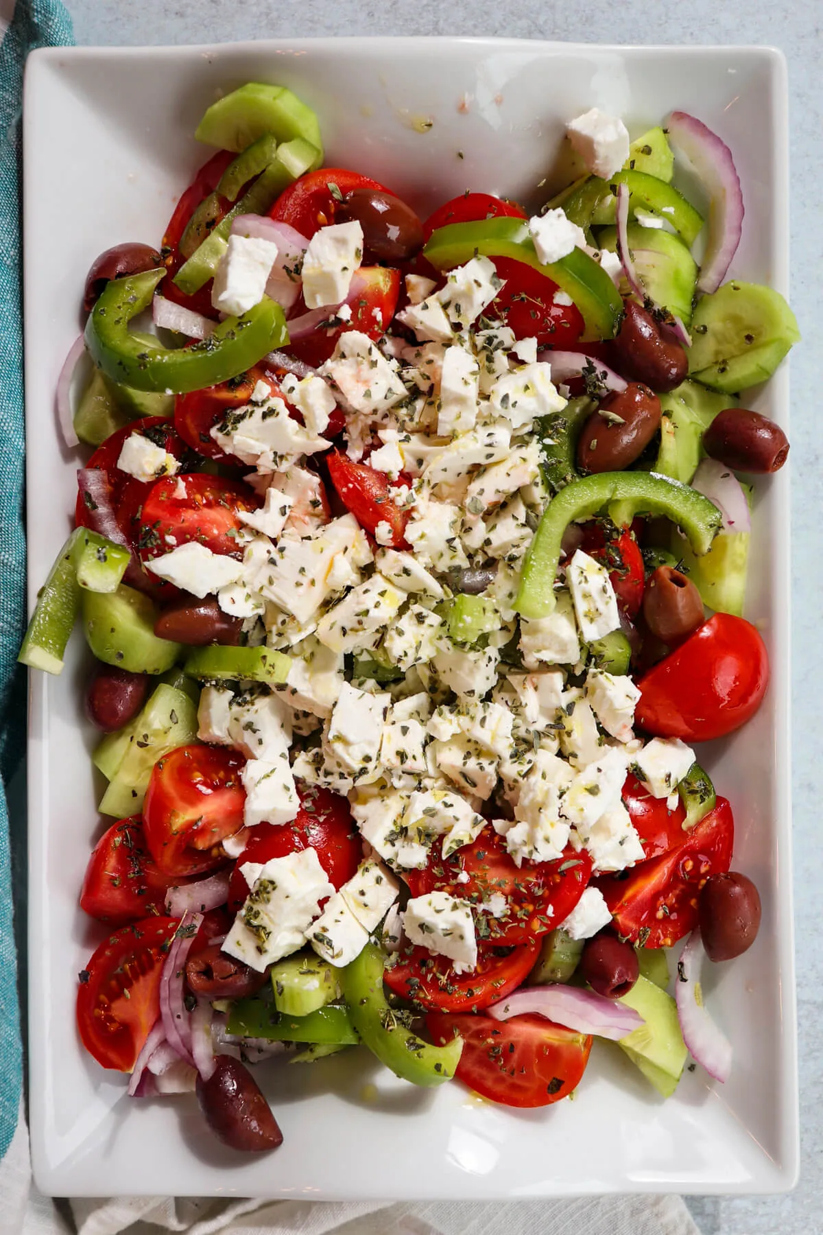 Overhead view of traditional Greek cucumber salad on a white serving dish with tomatoes, onions, olives, etc. and topped with crumbled feta cheese.