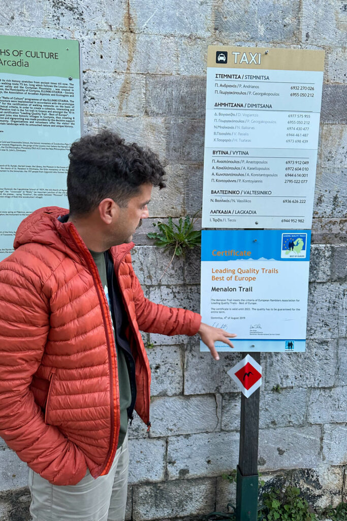 Alexandros in an orange jacket pointing to a Menalon Trail sign
