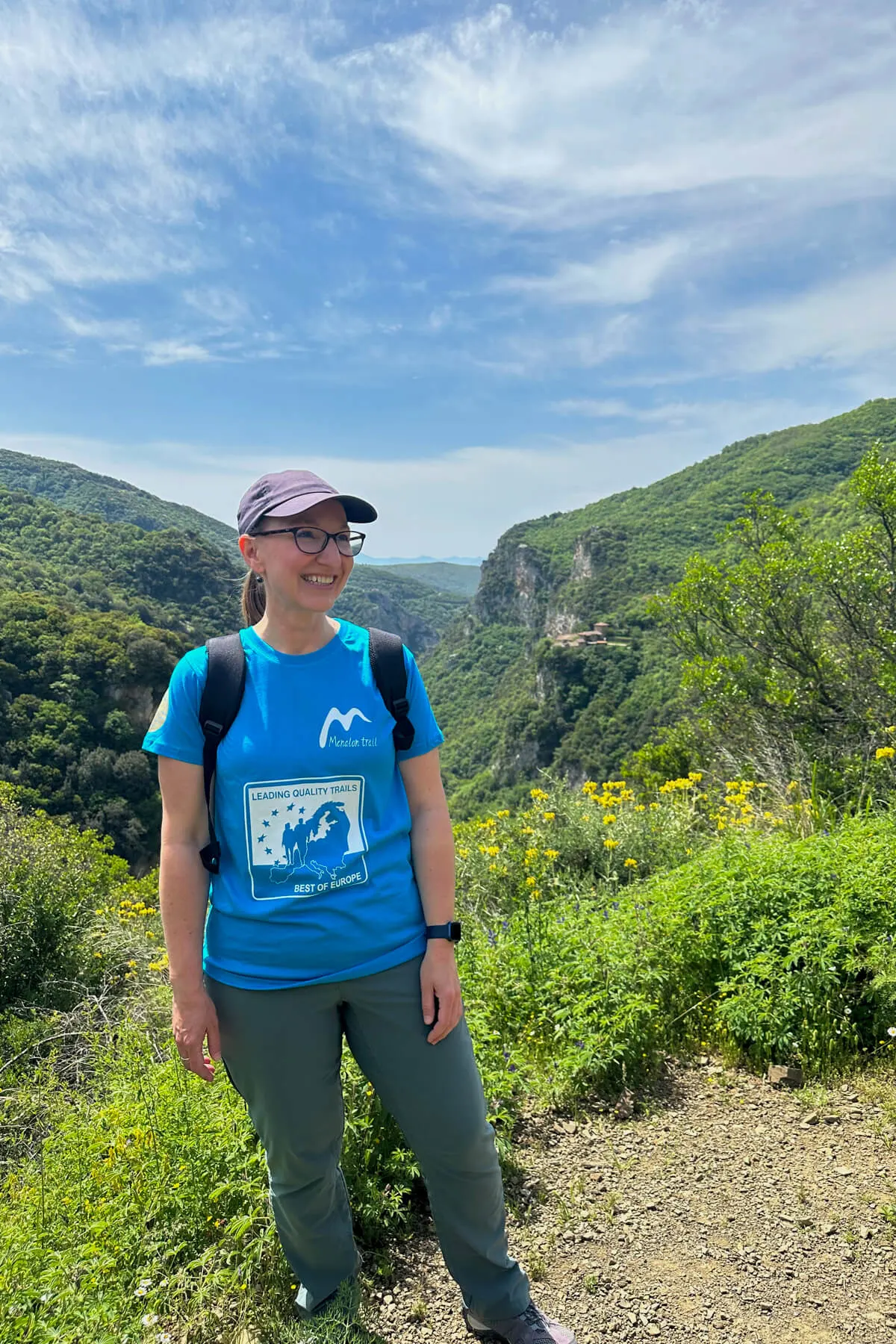 Lauren in a blue Menalon Trail tshirt and navy cap smiling in front of an overlook out to the Lousios Gorge, section 1 of the Menalon Trail