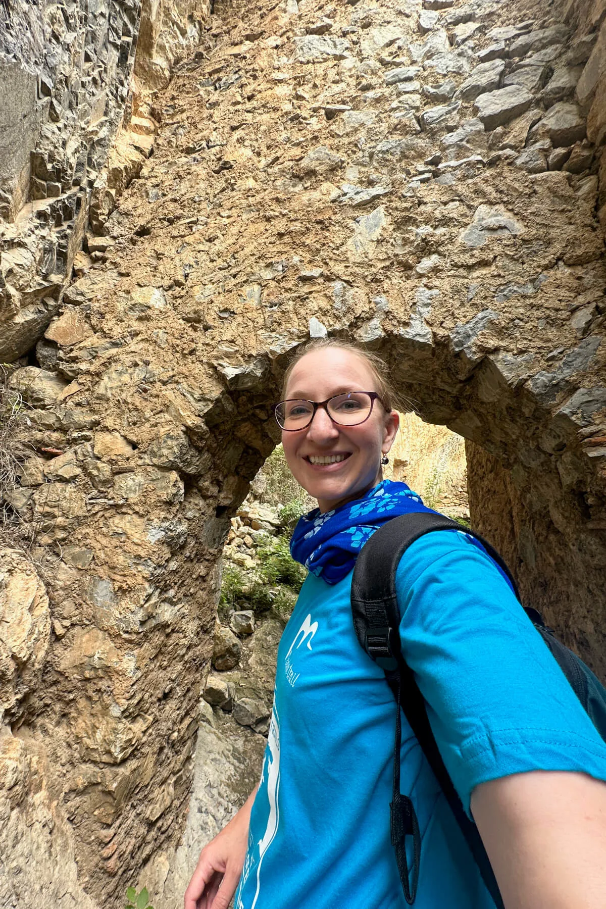 Lauren in a blue tshirt in front of the ruins of an old monastery with a stone archway between the wall and cliff face