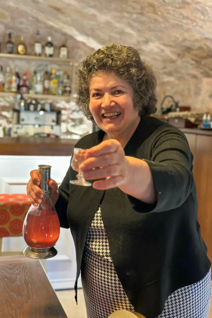 A hostess smiling in a black sweater welcoming us to her hotel holding out a small glass of homemade liqueur and Greek cookies and treats