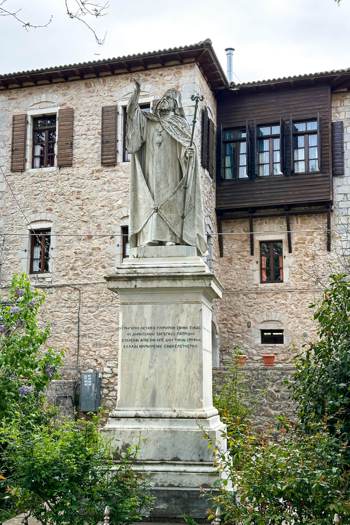 Statue of a bishop in Dimitsana in front of some brown buildings with shutters.
