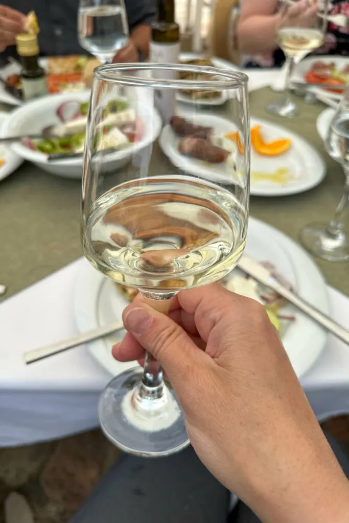 Glass of white wine in front of plates of Greek salad, sausages, etc.