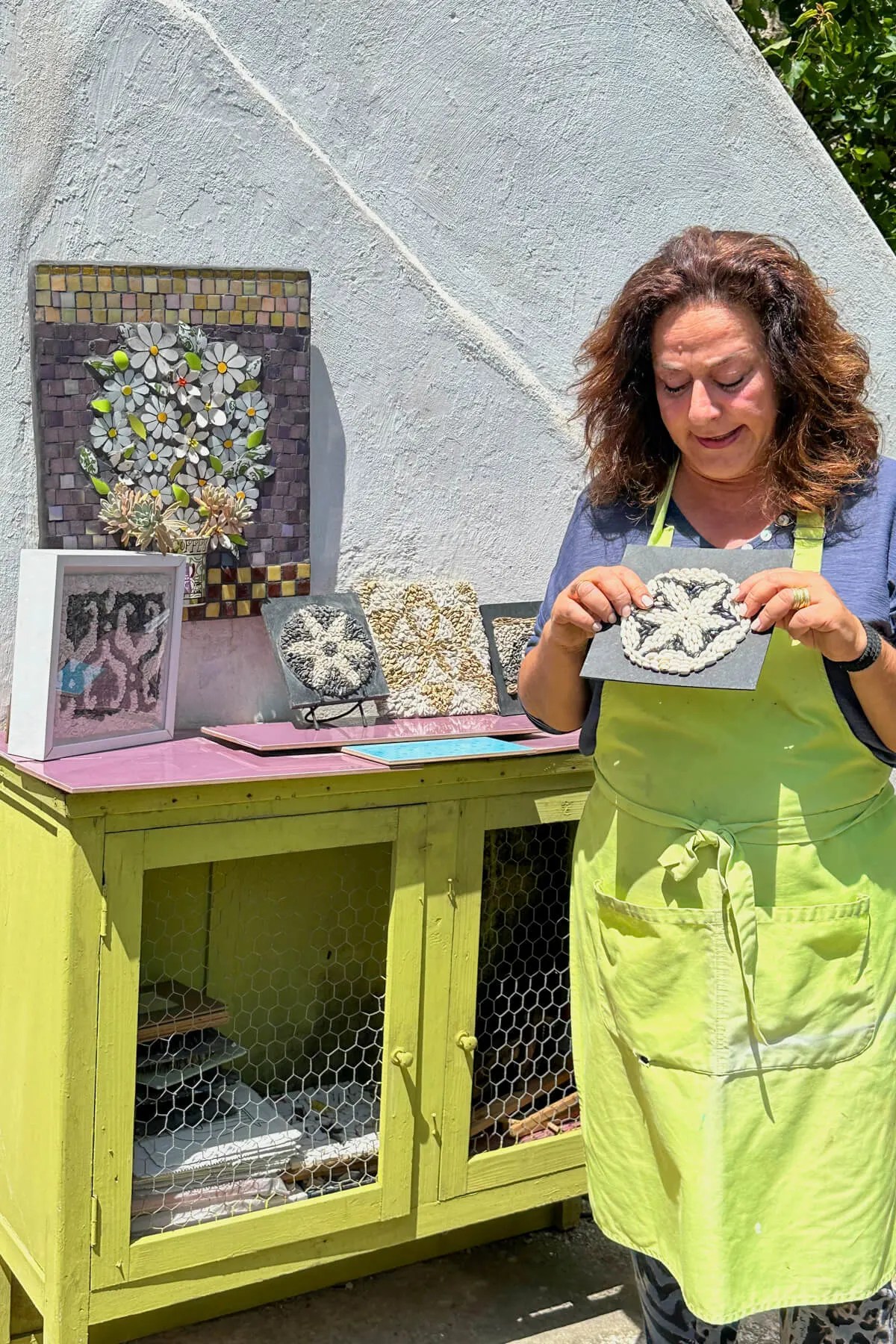 Dimitra demonstrating one of the techniques used in mosaics in a neon green apron next to a table with samples