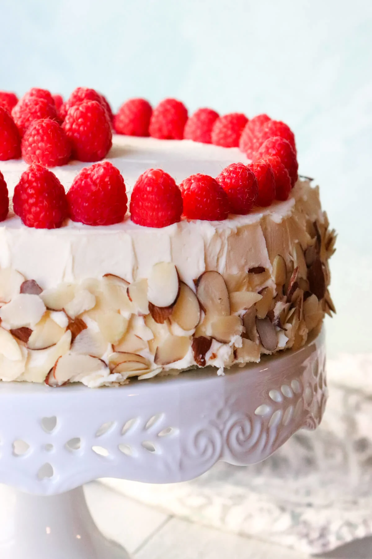 Side view of the keto almond raspberry cake on a white pedestal, decorated with fresh raspberries and almonds