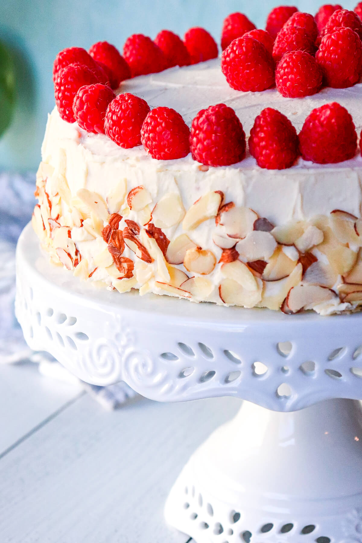 Side view of the keto almond raspberry cake on a white pedestal, decorated with fresh raspberries and almonds