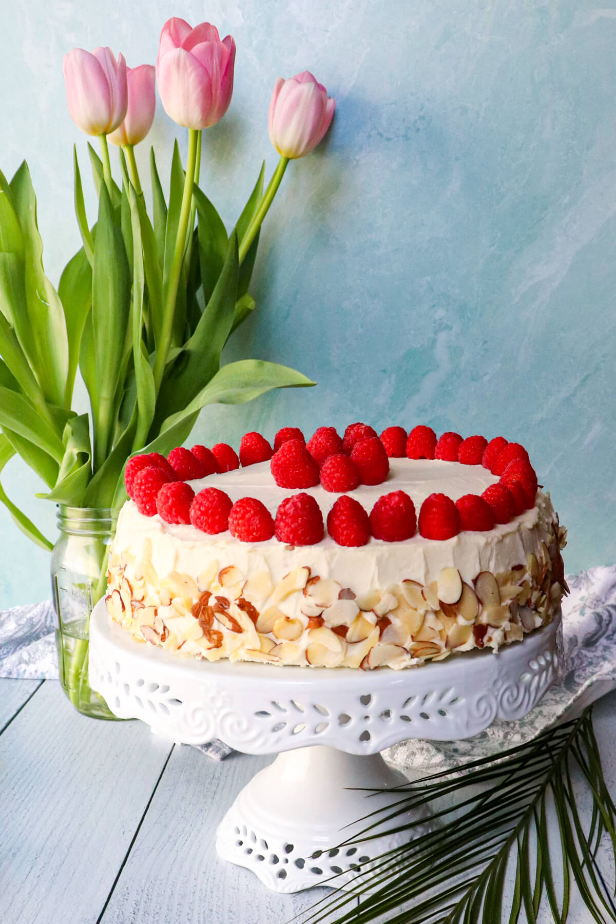 The almond raspberry keto cake box mix recipe cake on a white pedestal with pink tulips in the background