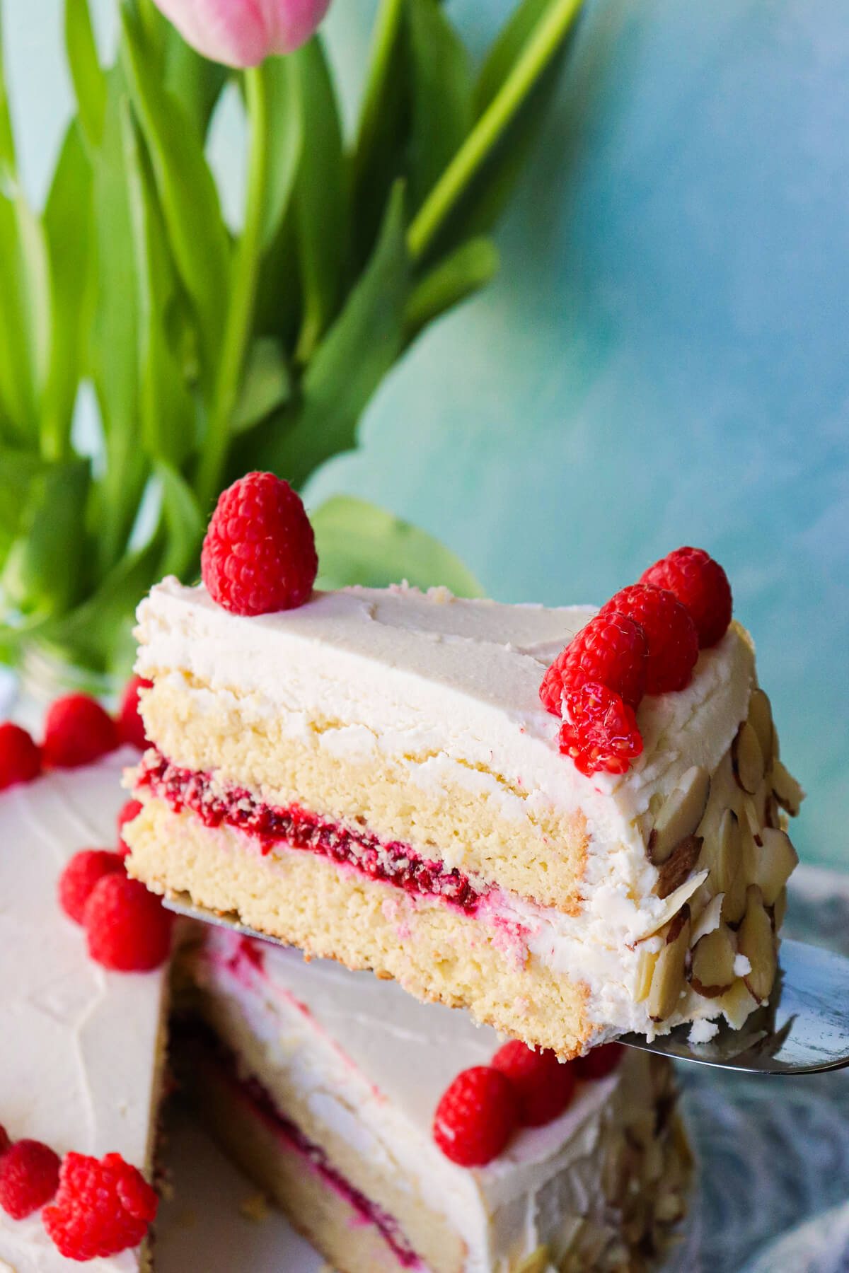 A keto almond raspberry cake with a keto cake box mix option on a white plate, with a slice being taken out of the entire cake dotted in raspberries