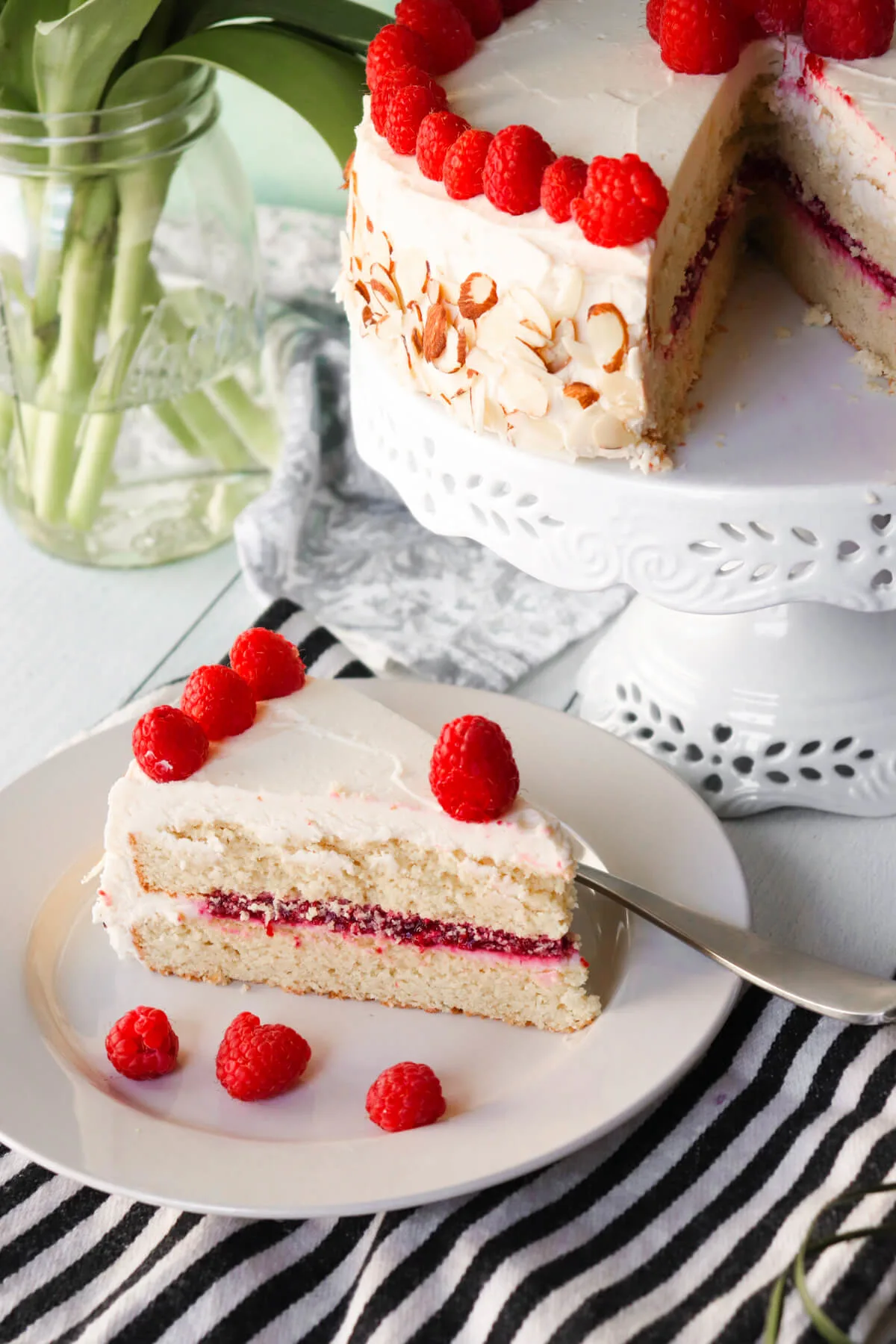 A keto almond raspberry cake with a keto cake box mix option on a white plate, with a slice taken out of the entire cake dotted in raspberries