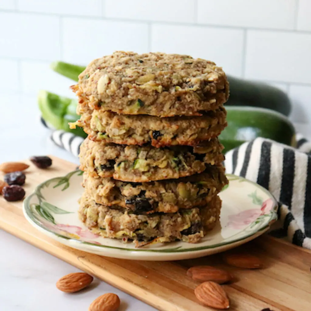 Gluten free zucchini breakfast cookies in a stack on a desert rose plate with zucchini in the background and almonds on the counter in front