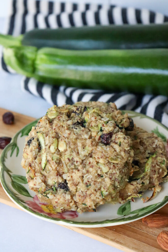 Two gluten free keto zucchini breakfast cookies a desert rose plate with zucchini in the background and almonds on the counter