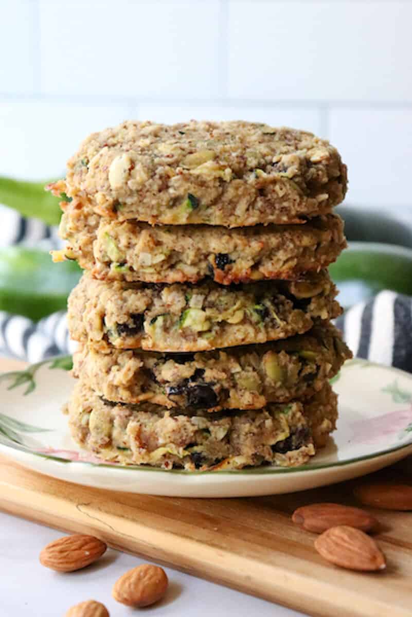 Gluten free zucchini breakfast cookies in a stack on a desert rose plate with zucchini in the background and almonds on the counter