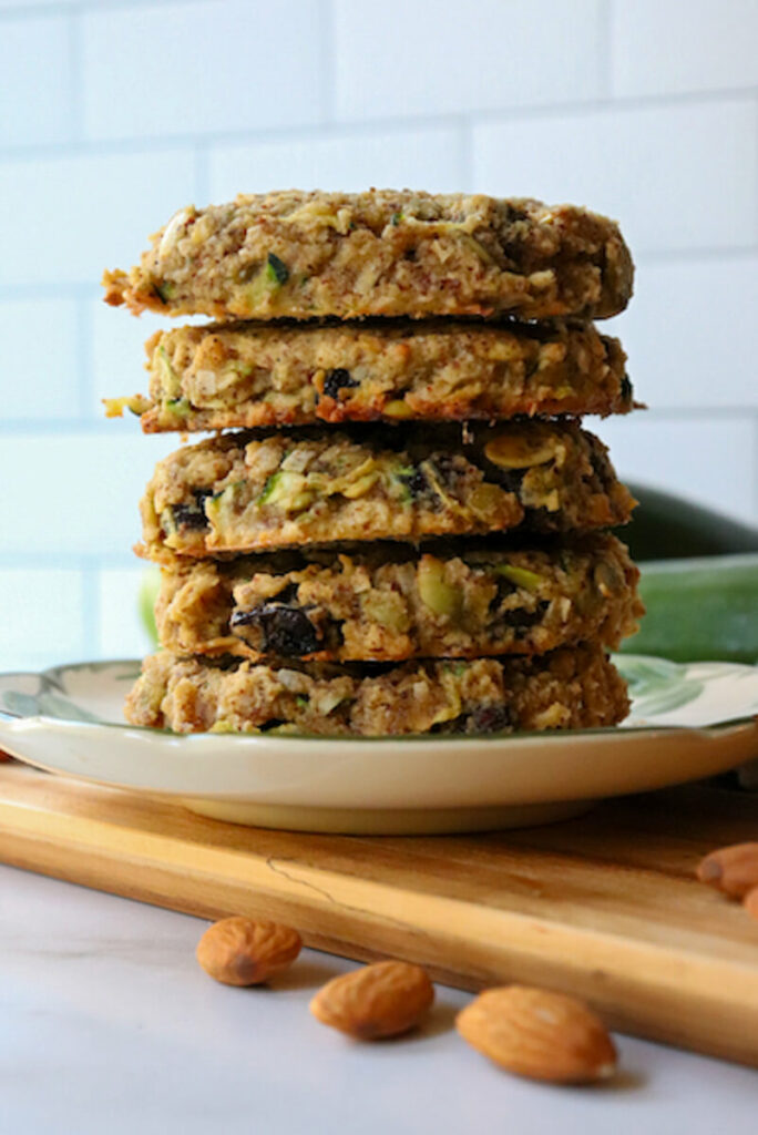 Gluten free zucchini breakfast cookies in a stack on a desert rose plate with zucchini in the background and almonds on the counter
