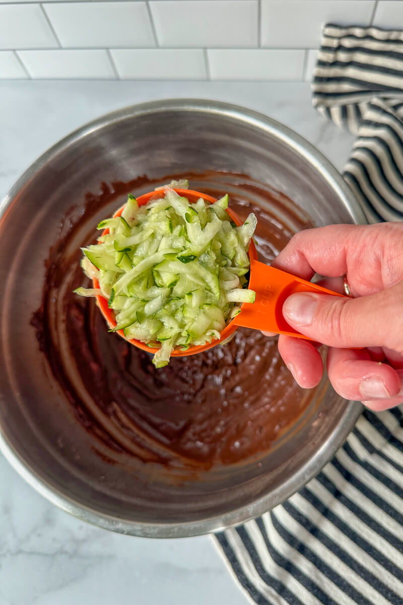 Bowl of keto zucchini brownie batter with a cup of grated zucchini over it.