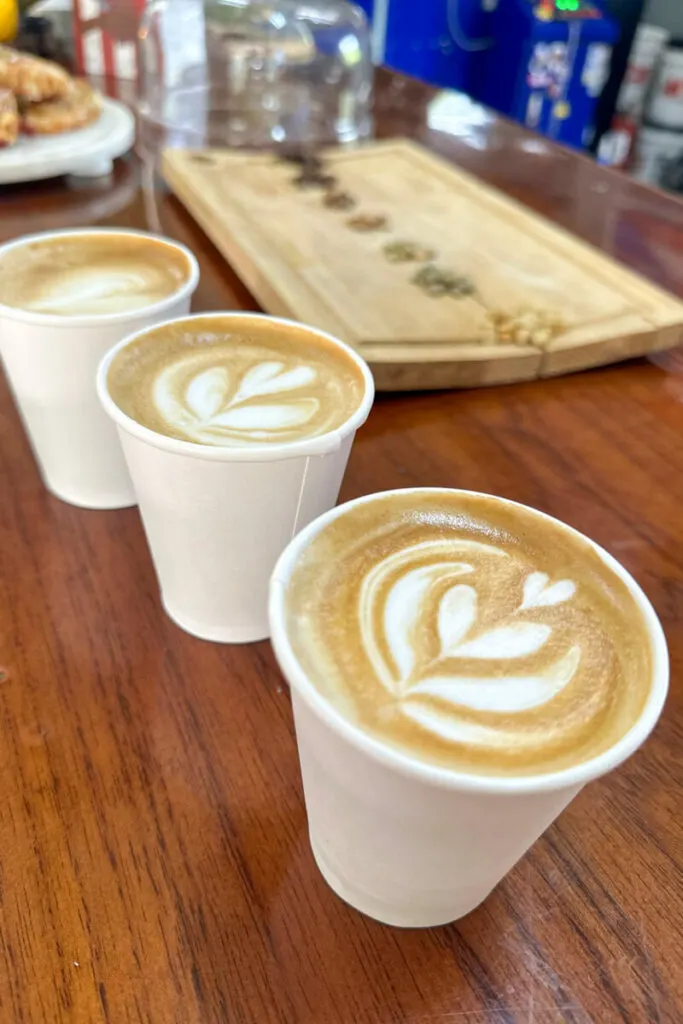 Three cups of coffee in front of a wooden display of different coffee bean roasts