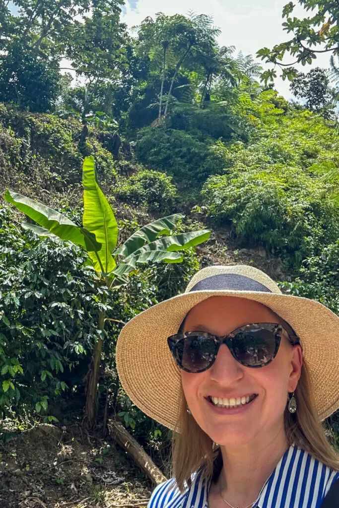 A woman in a white hat in front of a dense forest and hill with a banana tree