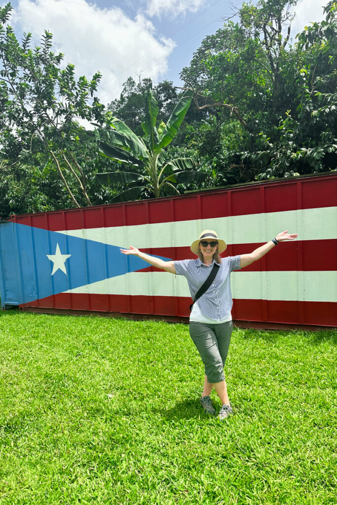 A woman in a white hat and capris standing with arms raised in the grass in front of a Puerto Rican flag