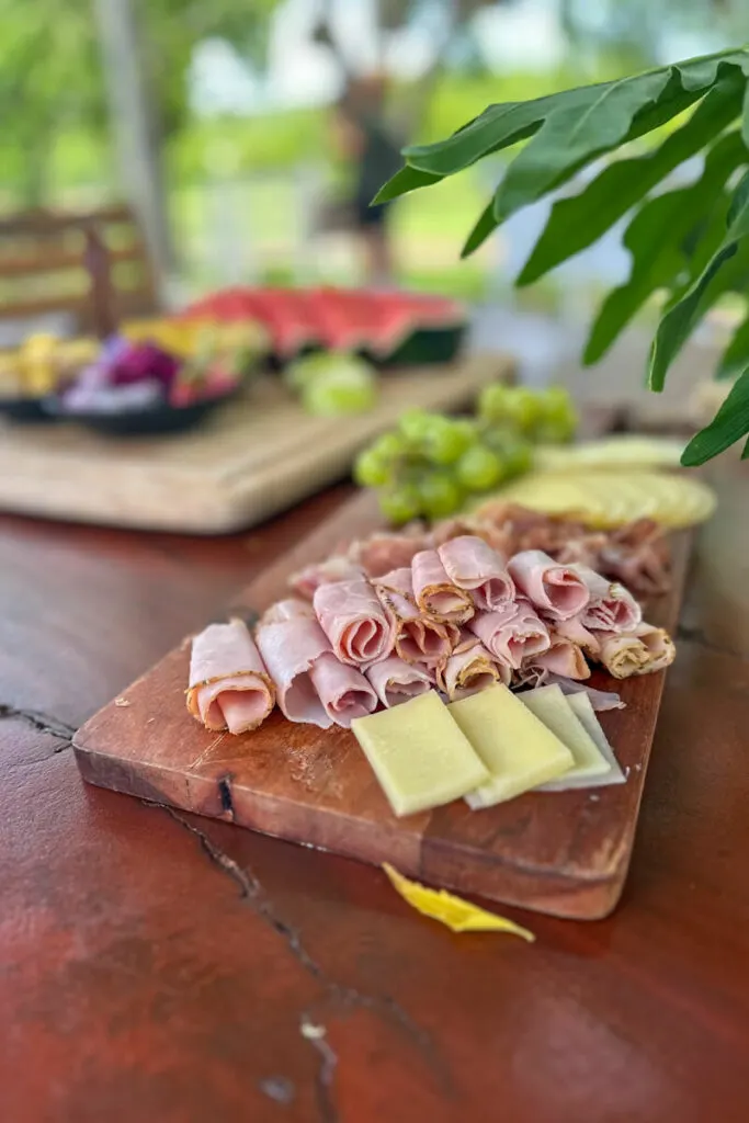 A wooden table with a wooden cutting board with deli meat, cheese, and grapes. There is more fruit in the background.