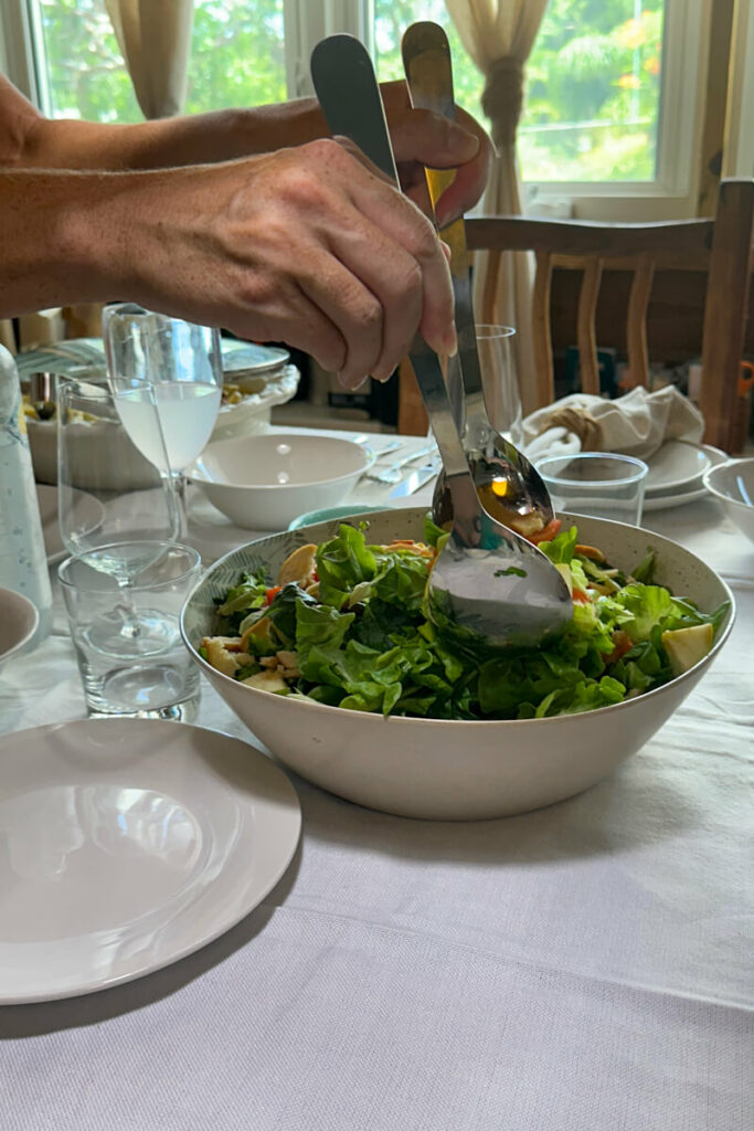 A white table clothe covered table with a view of two hands getting salad out of a bowl with spoons