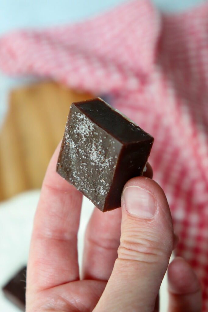 A hand holding up a piece of protein keto fudge in front of a red and white towel and wooden cutting board