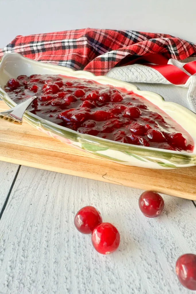 Boat style dish filled with homemade rich red cranberry sauce on a wooden platter with cranberries around.