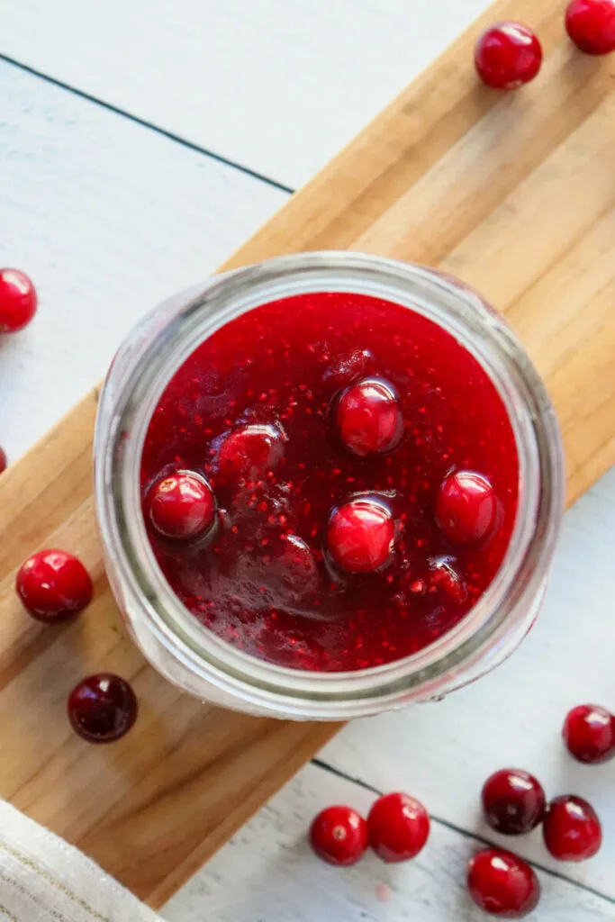 Overhead view of a mason jar of keto cranberry sauce on a wooden platter surrounded by cranberries.