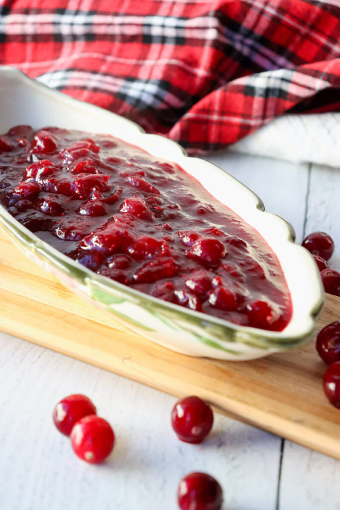 Boat style dish filled with homemade rich red cranberry sauce on a wooden platter with cranberries around.