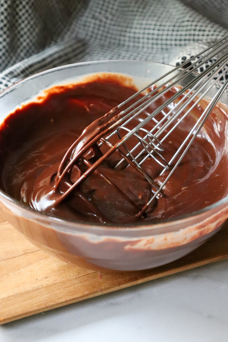 A clear glass bowl of keto chocolate ganache with a whisk stirring it.
