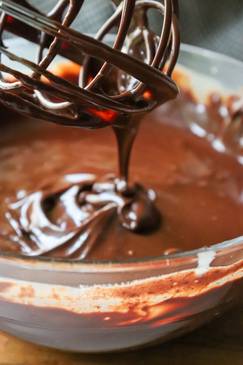 A clear glass bowl of keto chocolate ganache with a whisk stirring it and some dripping back down into the bowl.