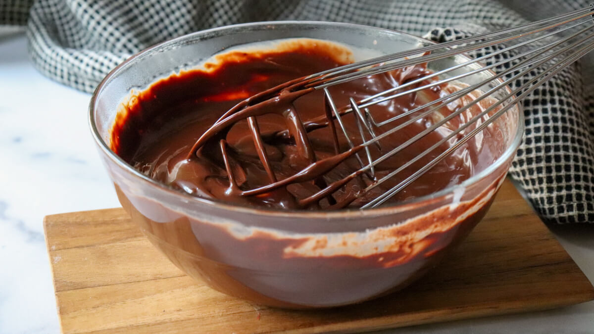 A clear glass bowl of keto chocolate ganache with a whisk stirring it.