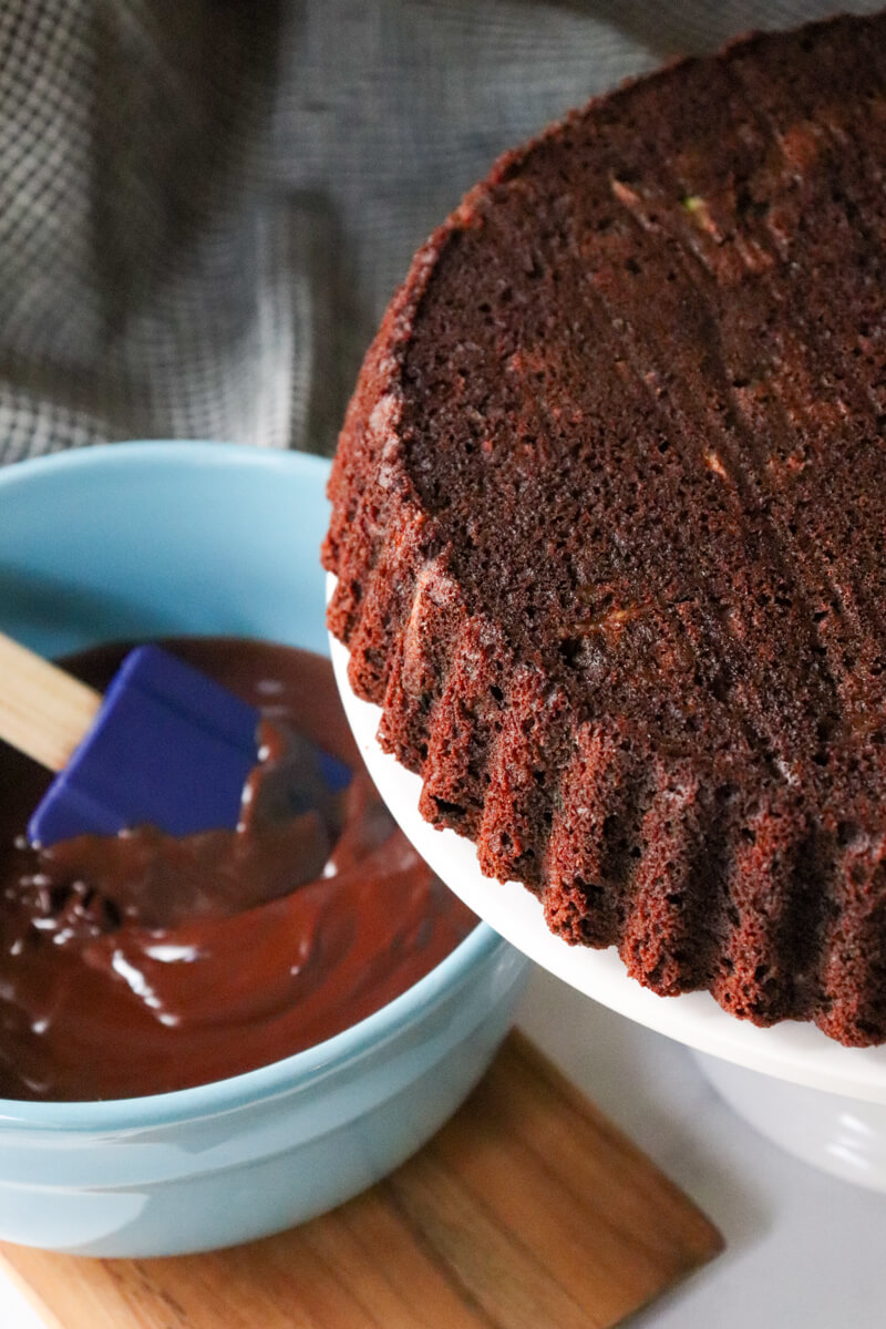 A keto chocolate zucchini cake on a pedestal with a blue bowl of keto chocolate ganache in the background.