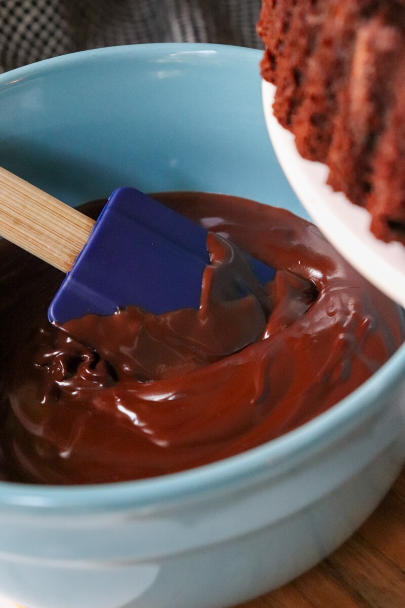 A keto chocolate zucchini cake on a pedestal with a blue bowl of keto chocolate ganache in the background.