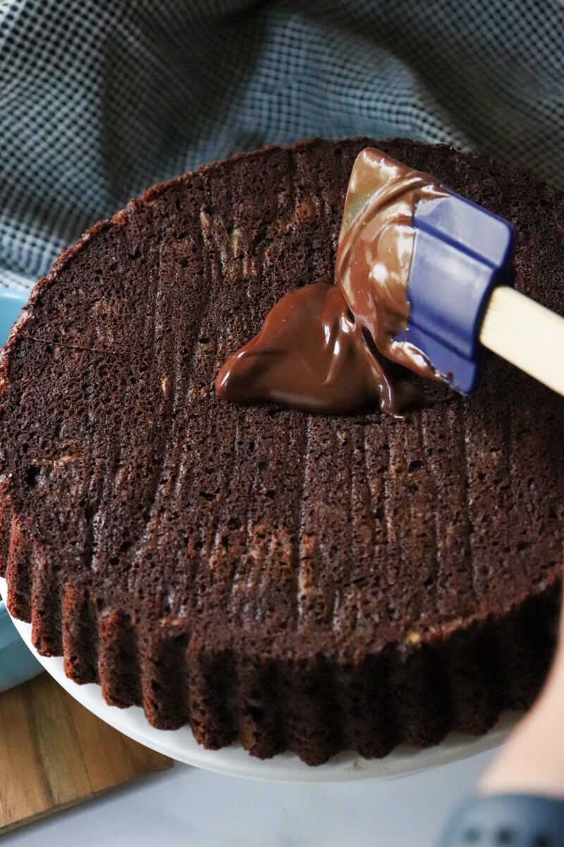 A keto chocolate zucchini cake on a pedestal with a blue spatula spooning the ganache on the low carb zucchini cake.