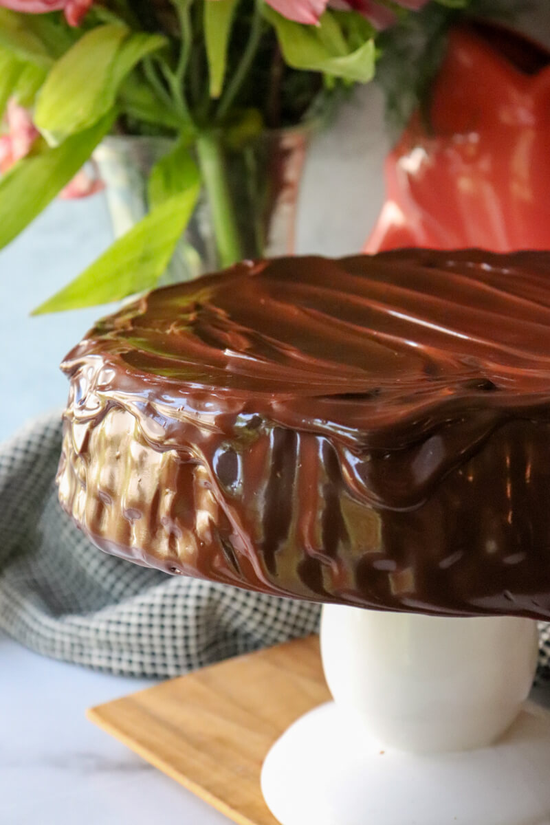 Front view of low carb zucchini chocolate cake with keto chocolate ganache on a white pedestal. In front of greenery.