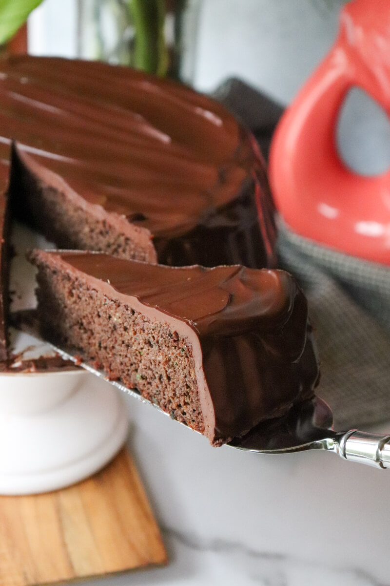 A slice of keto zucchini chocolate cake with low carb ganache on a server being pulled out of the cake on a white pedestal.