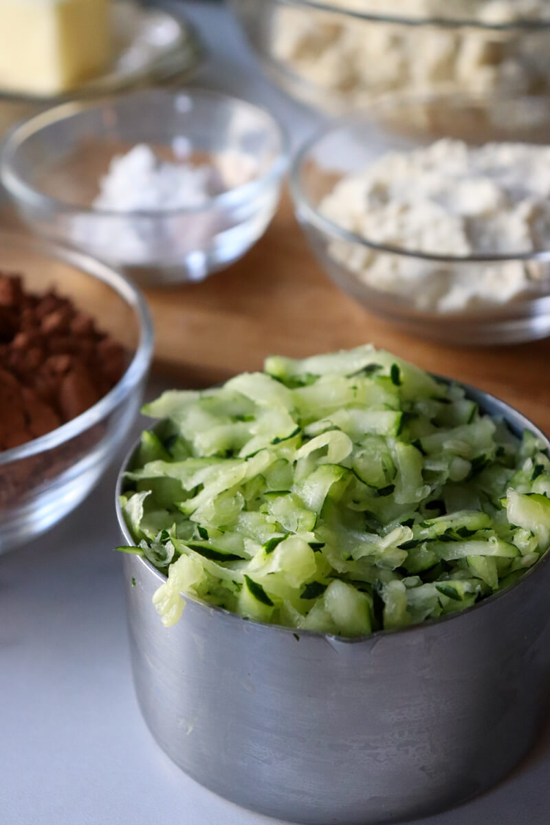 Front view of the grated zucchini in a metal measuring cup with other ingredients in the background.