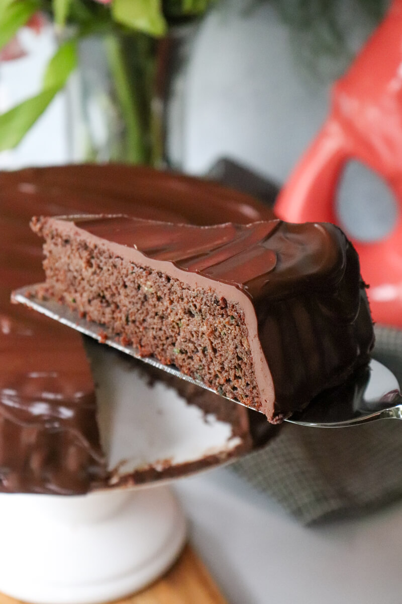 A slice of keto zucchini chocolate cake with low carb ganache on a server being pulled out of the cake on a white pedestal.